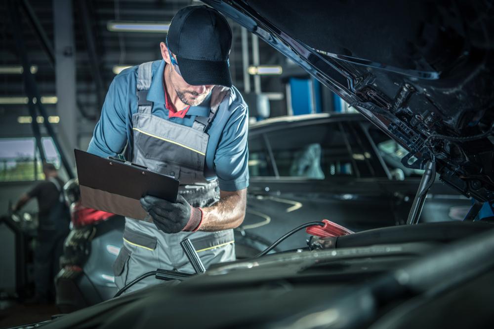 Man in overalls and a baseball cap, looking under the hood of a car and holding a clipboard