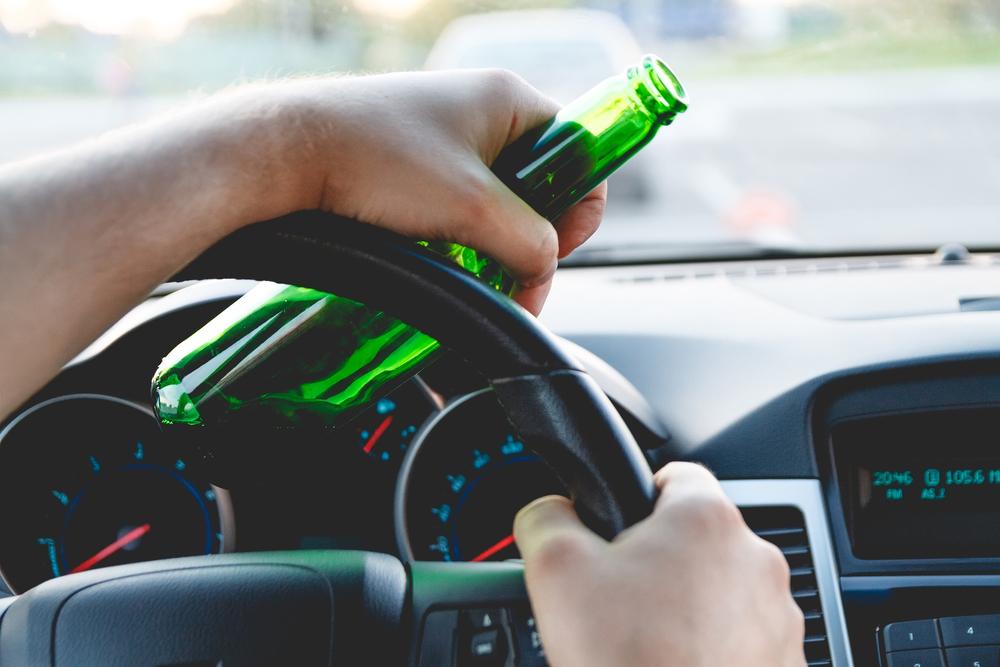 Car dashboard, one hand on the steering wheel and another holding a beer bottle