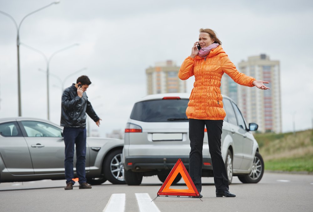 A woman is frustrated as she talks on the phone next to a car wreck.