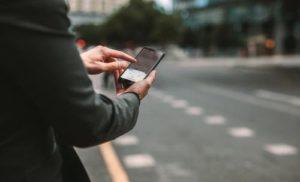 A man standing next to the street holding a smartphone and navigating a rideshare app